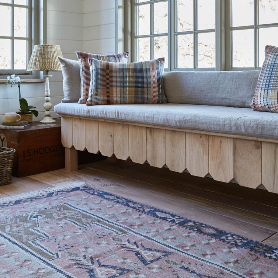 An Aster rug with a geometric pattern in pink, green, and gray colors, laid out on a wooden floor in a room with a sofa and window in the background.