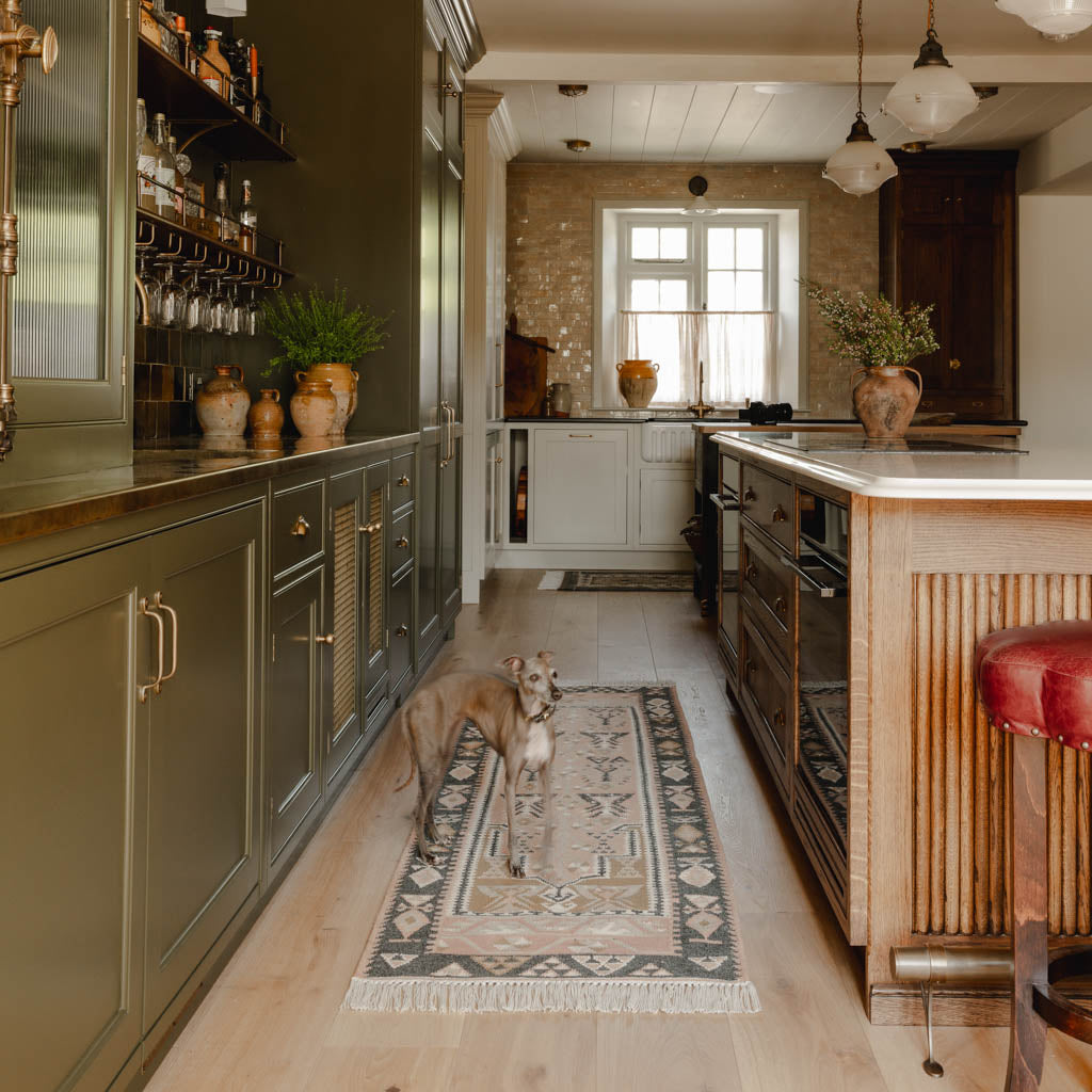 Dog standing on a patterned rug in a modern kitchen with wooden island and green cabinets.