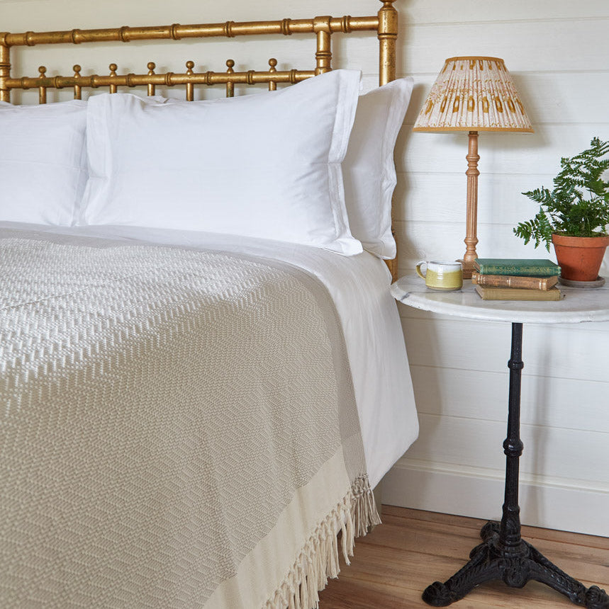 A grey chevron throw blanket draped over the end of a bed, with a bedside lamp and plant on the side table.
