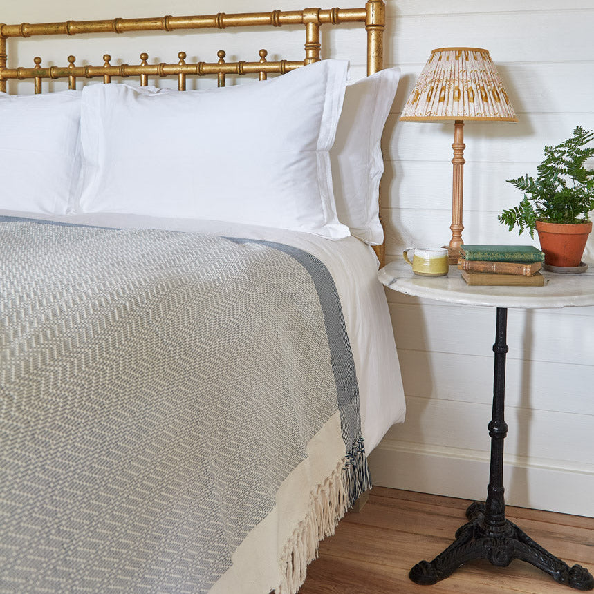 A navy chevron throw blanket with fringed edges is draped over the edge of a bed, with a bedside table and lamp visible in the background.