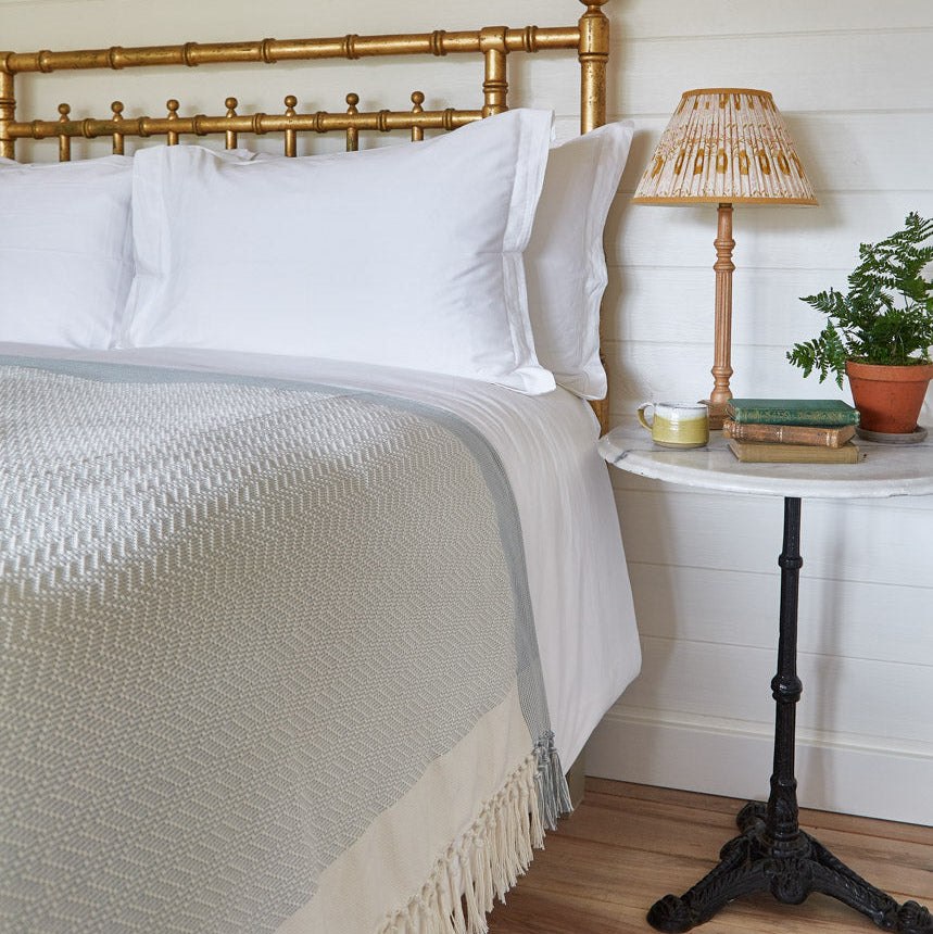 A sky blue chevron throw blanket with fringe detailing, draped over the edge of a bed, with a bedside lamp and plant in the background.