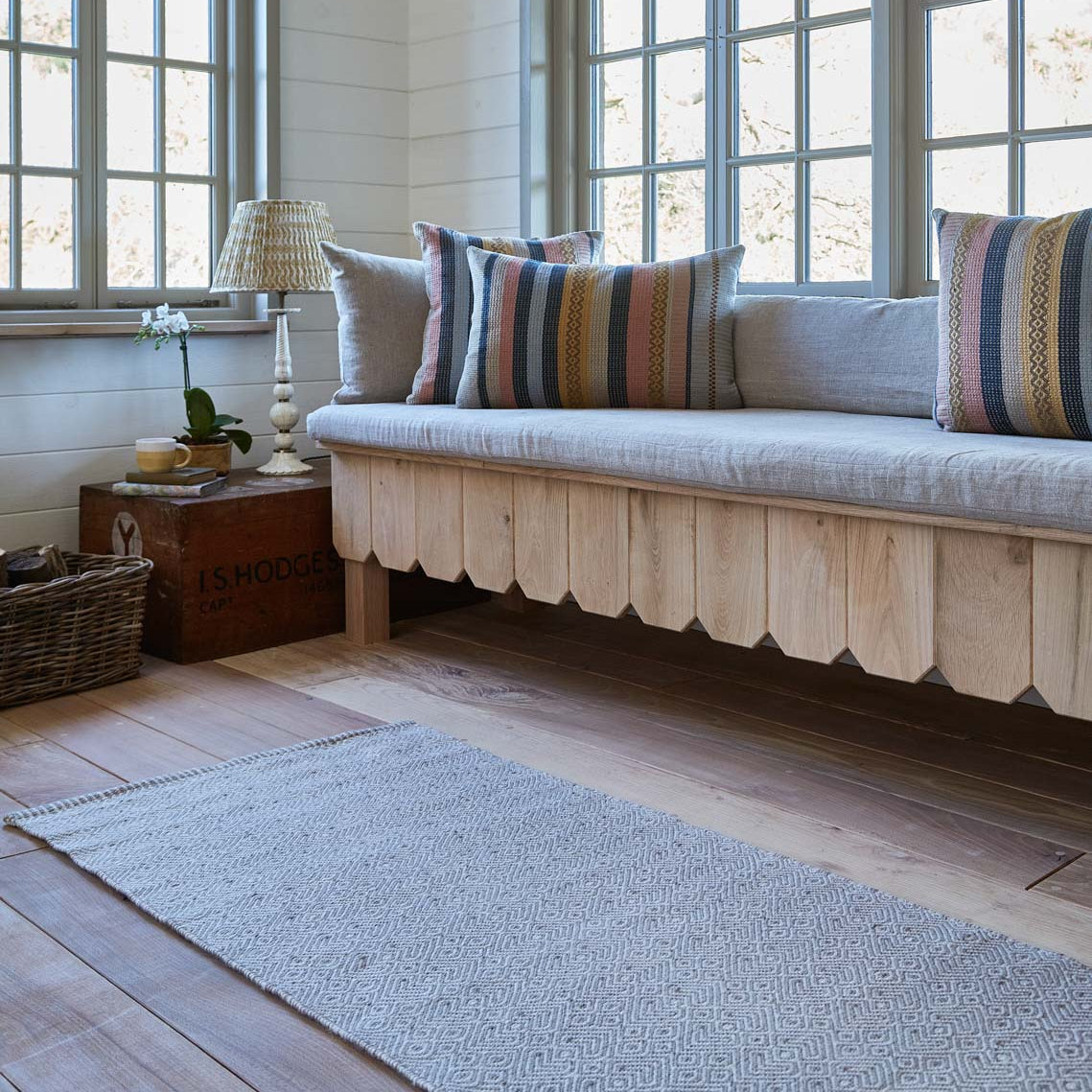 A grey runner rug made from recycled plastic bottles, laid out on a wooden floor in a room with a wooden bench and multicolored pillows.