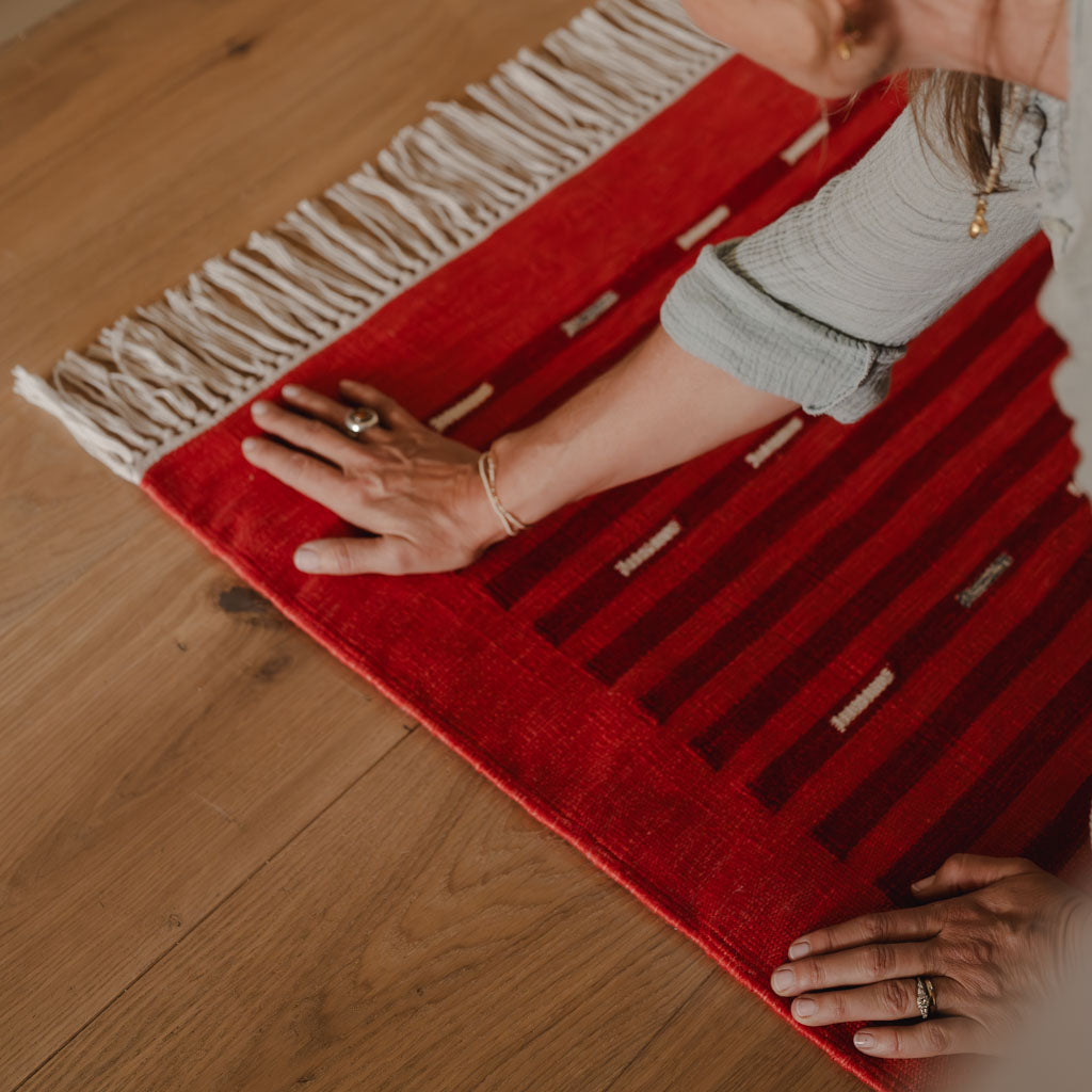 Person holding a red rug with white fringe on a wooden floor