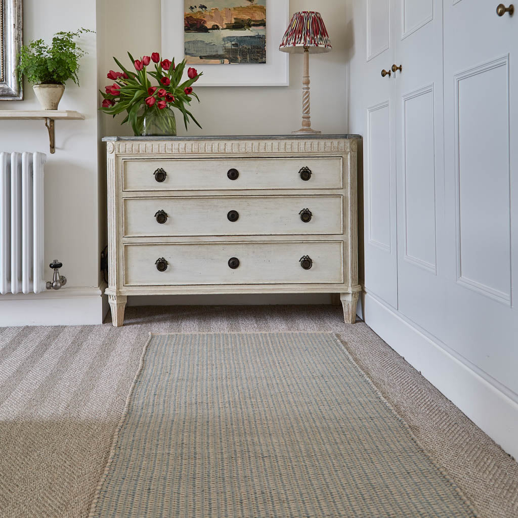 A striped celadon rug placed on the floor in front of a dresser, with a floral arrangement on the dresser and a lamp in the background.
