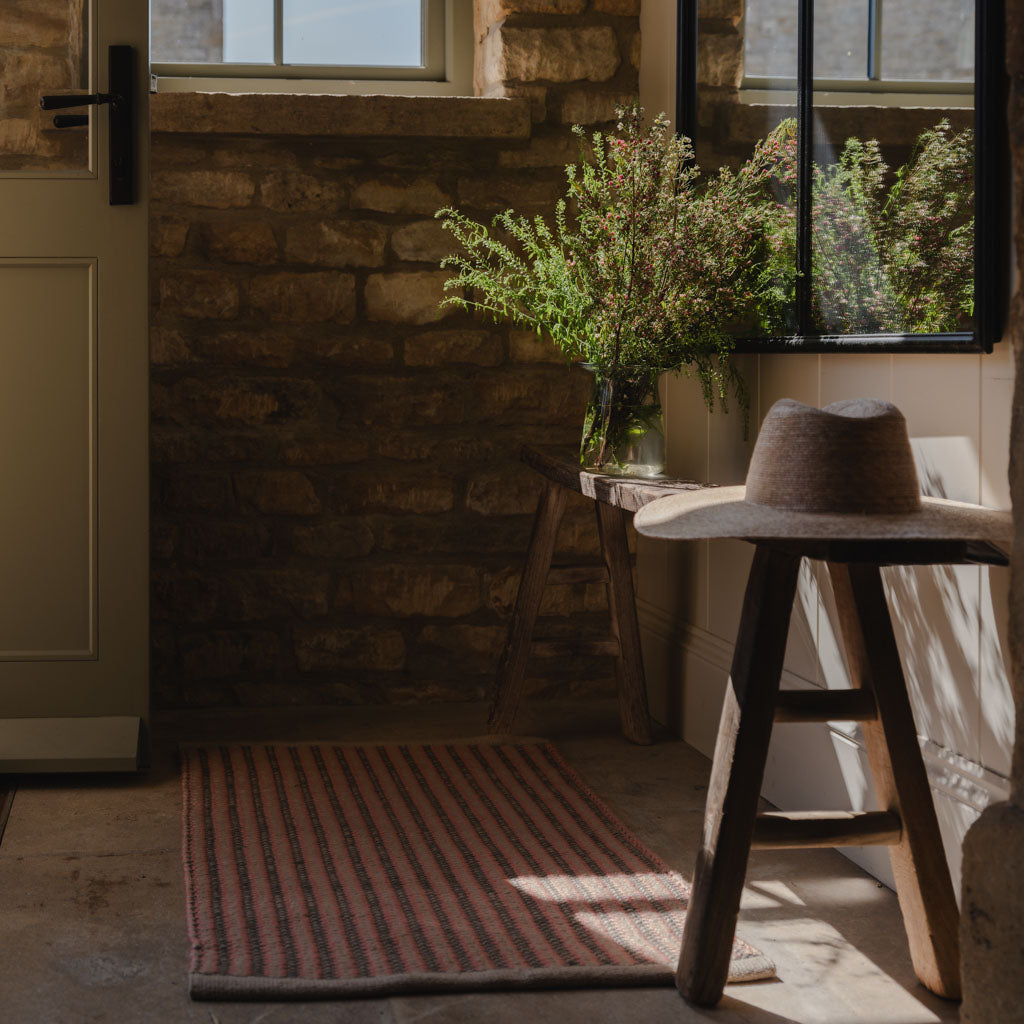 Nestled corner of a room with a wooden stool, striped coral kilim rug, and window view.