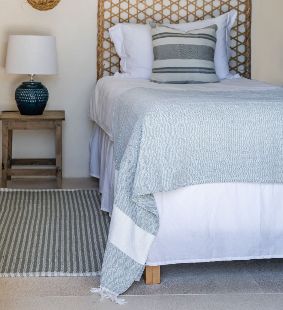 Bedroom with a bed covered in a light blue and white striped blanket, wooden nightstand with lamp, and woven headboard.
