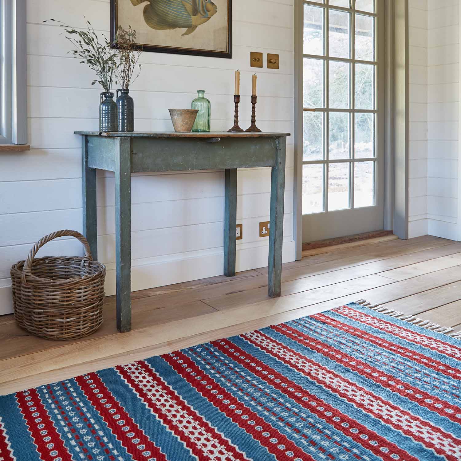 A red and blue striped rug with a geometric pattern laid out on a wooden floor, leading up to a rustic style table with a vase and other decorative items.