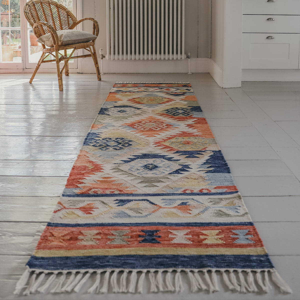 Native American inspired runner rug with motif repeat, in blues, greens, corals and neutrals on a wooden floor with a wicker chair and radiator in the background.