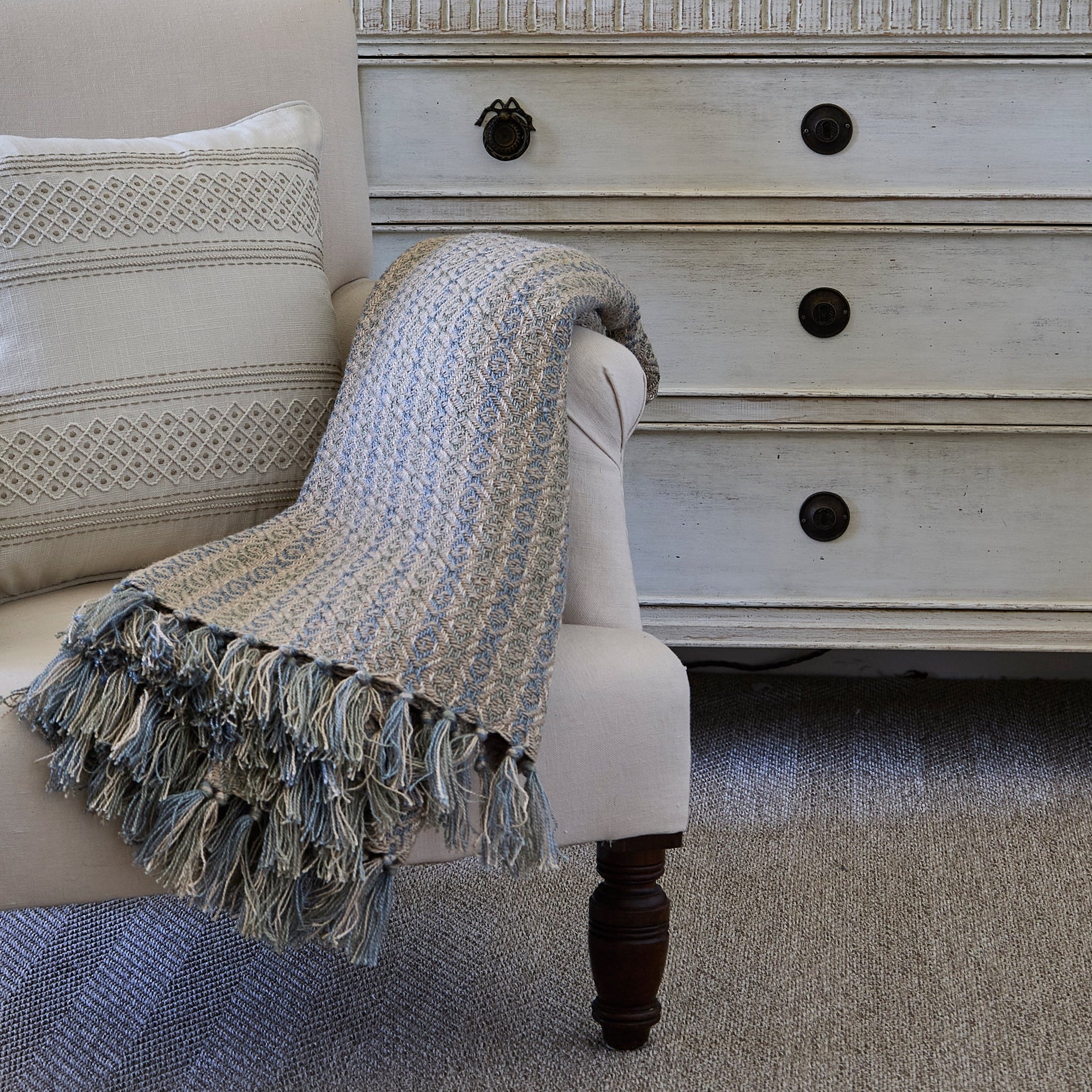 White armchair with a blanket and pillow in front of a white dresser with black knobs.