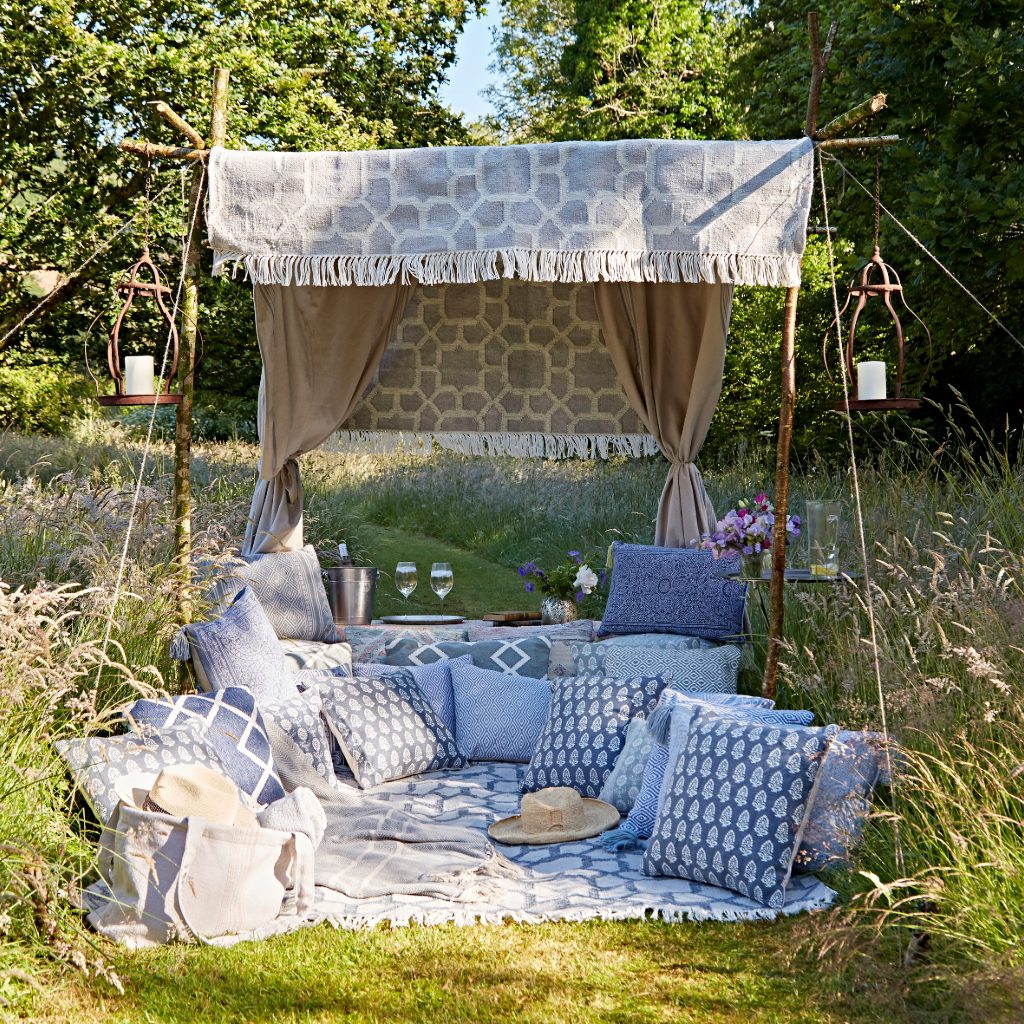 Outdoor seating area with blue and white patterned cushions, pillows, and a blanket under a canopy in a garden setting.