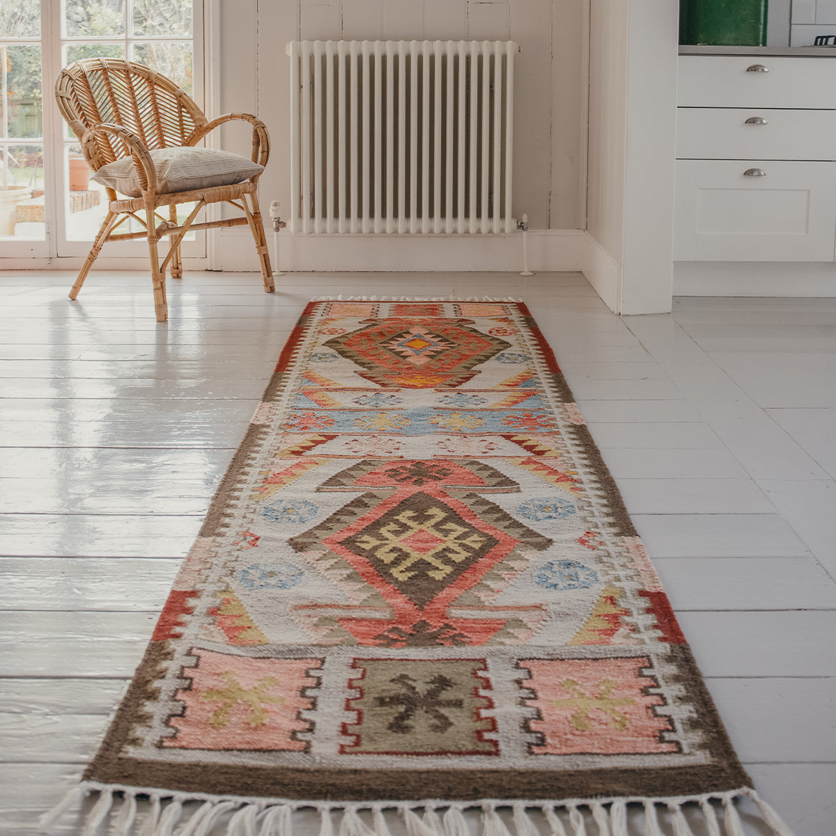 Runner rug with a geometric pattern in soft umber, burnt orange, faded blues and earthy saffron tones on a white wooden floor in a room with a chair and radiator.