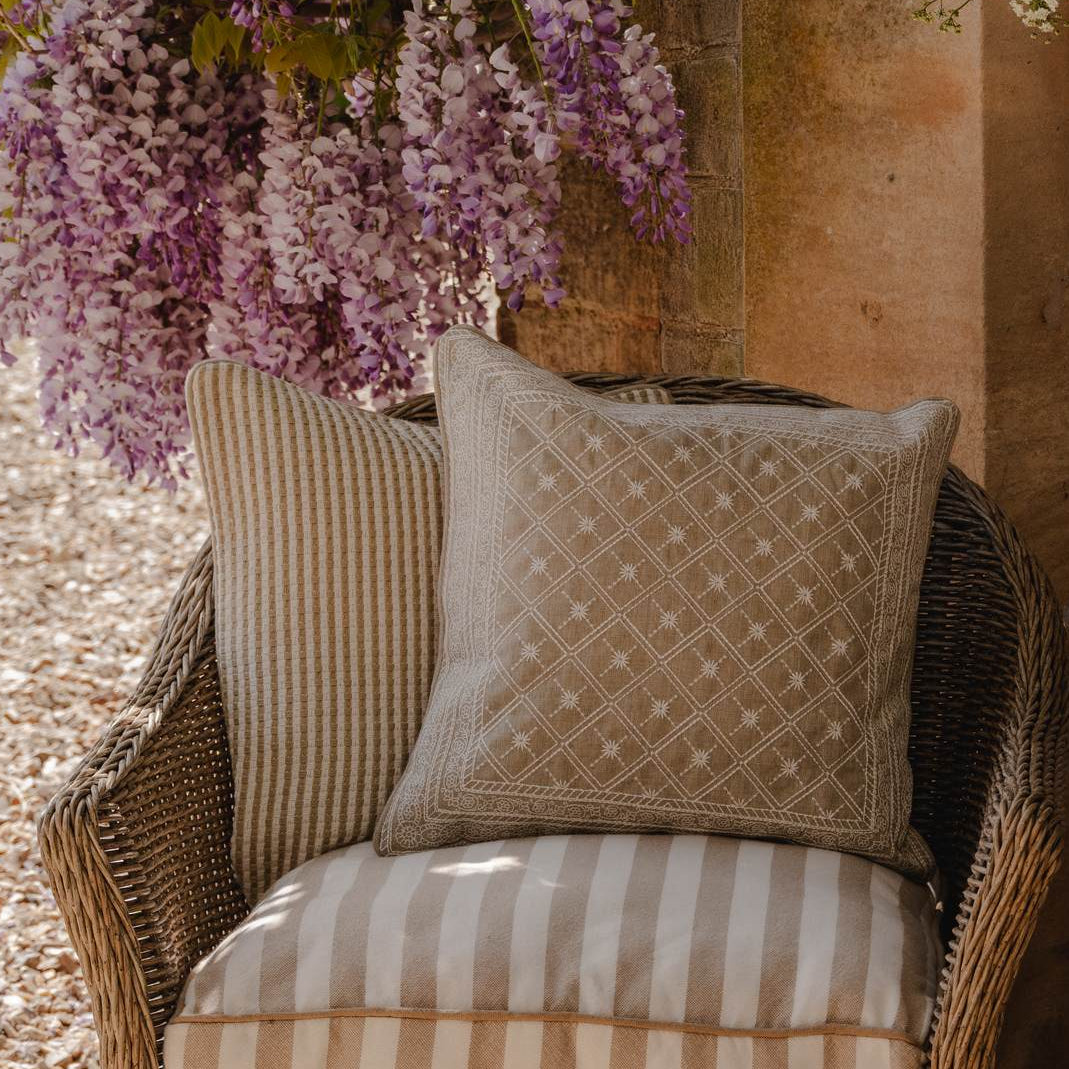 Wicker chair with embroidered cushions on a patio, surrounded by flowers and a building. *collection_outdoor