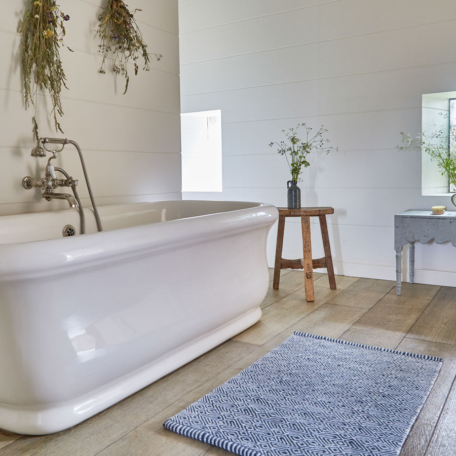 A navy blue and white geometric patterned rug placed on a wooden floor, beside a white bathtub and a small wooden stool, with a vase of flowers on the stool.