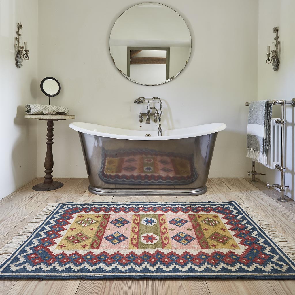 A multicolored geometric patterned rug with pink, blue, and yellow borders placed on a floor in front of a bathtub and a round mirror.