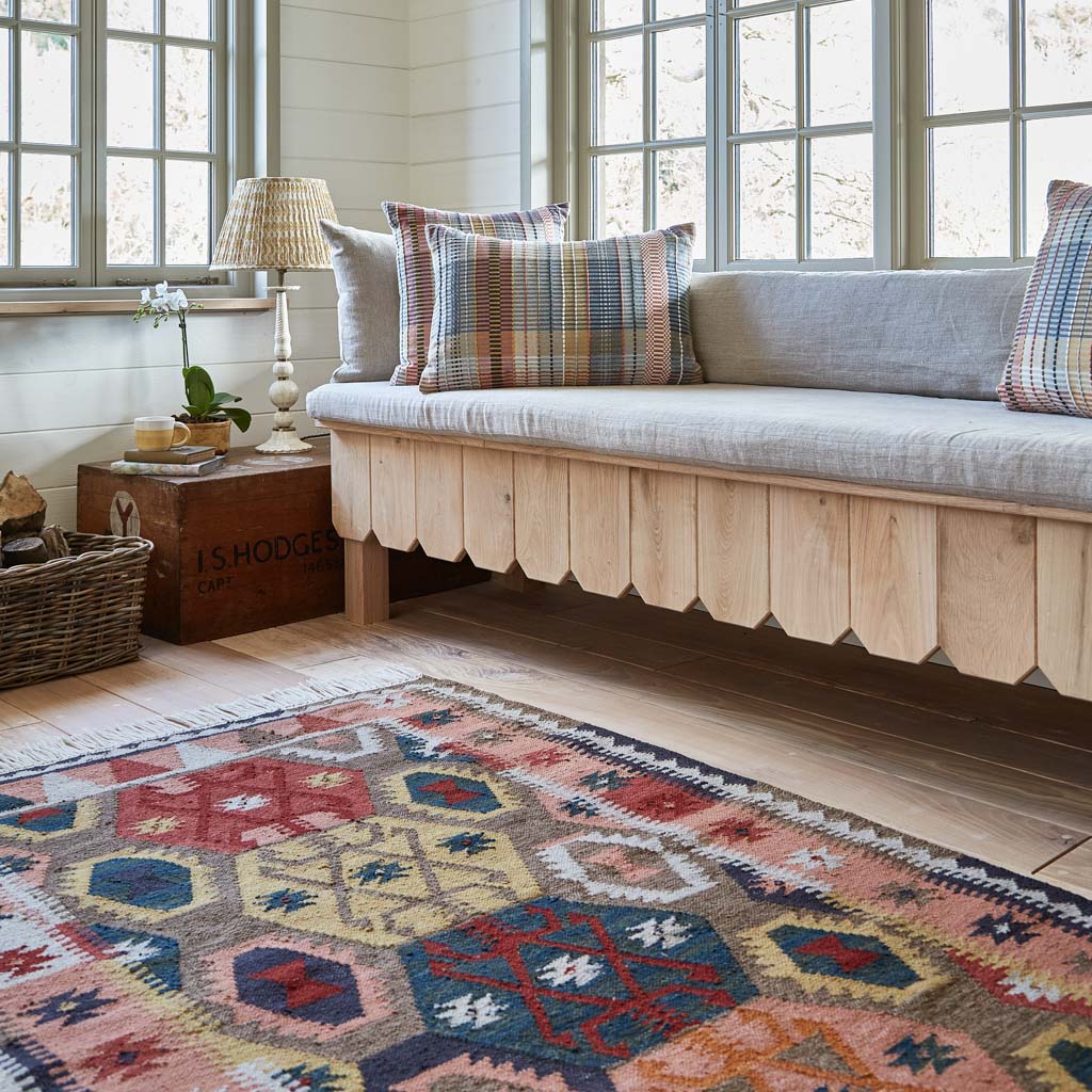 A multicolored geometric patterned rug with rich ochre and soft apricot hues, placed in a room with a bench and pillows.