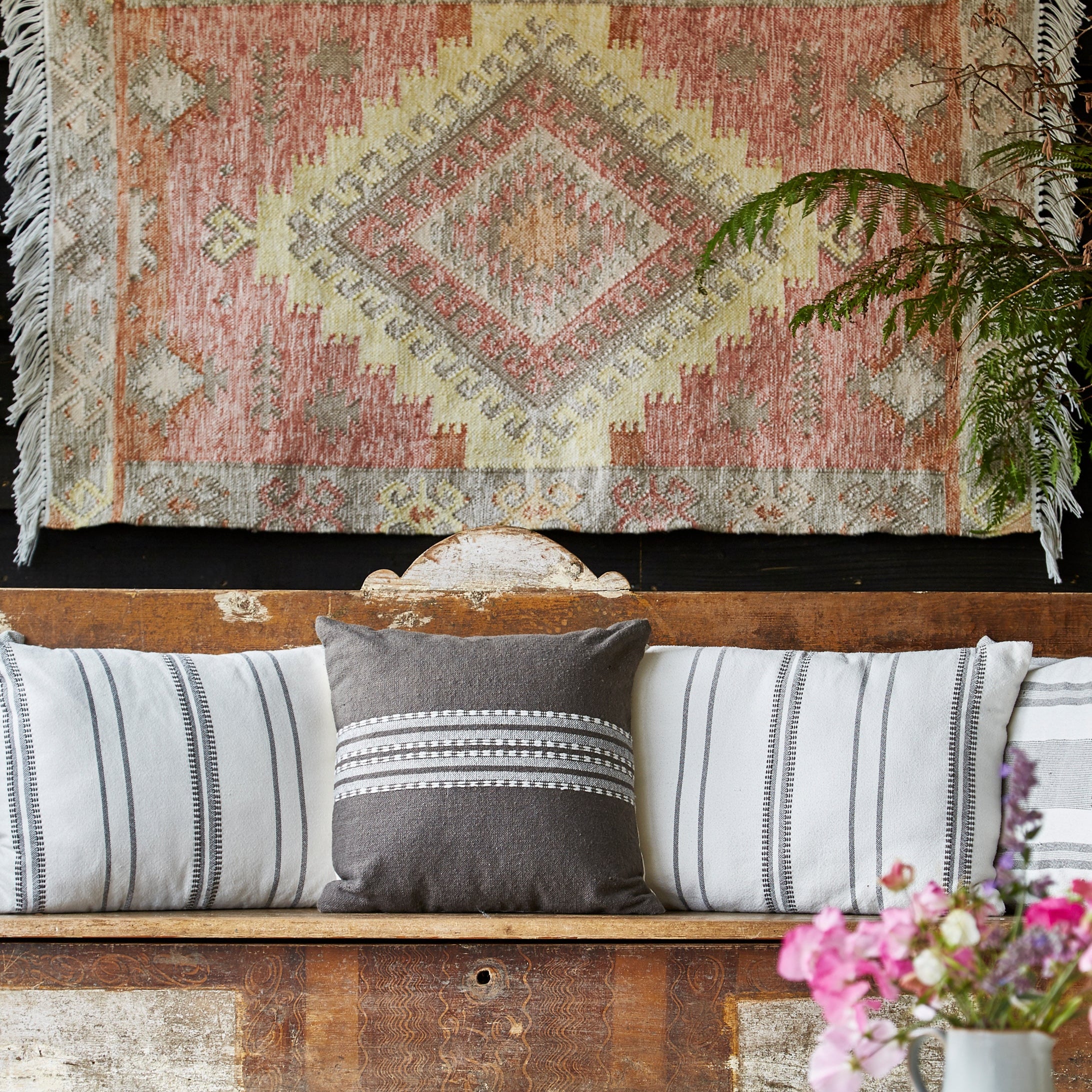 Decorative rug with geometric pattern on a wooden bench with striped cushions and a vase of flowers.