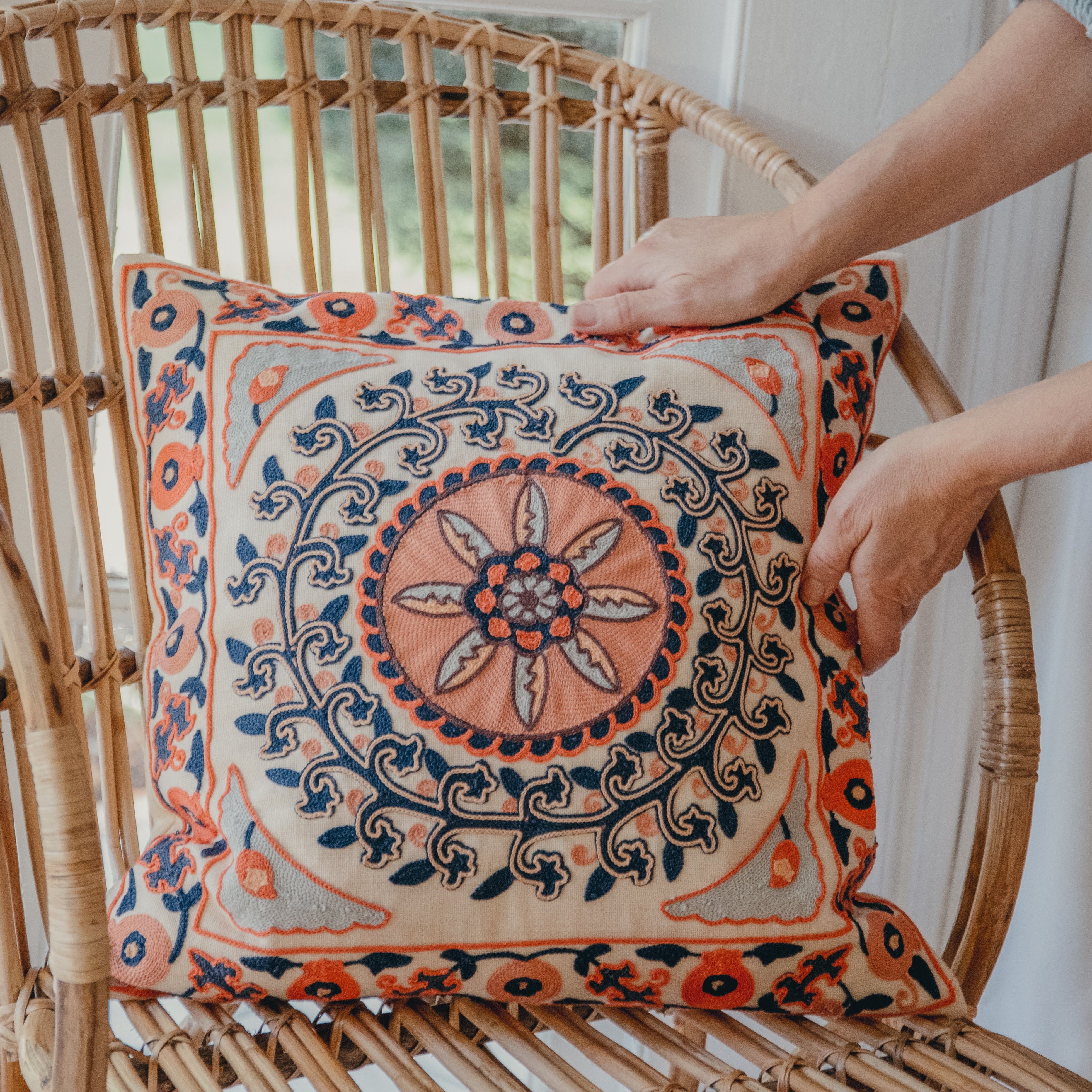 Embroidered cushion with coral and navy floral geometric pattern on a wicker chair.