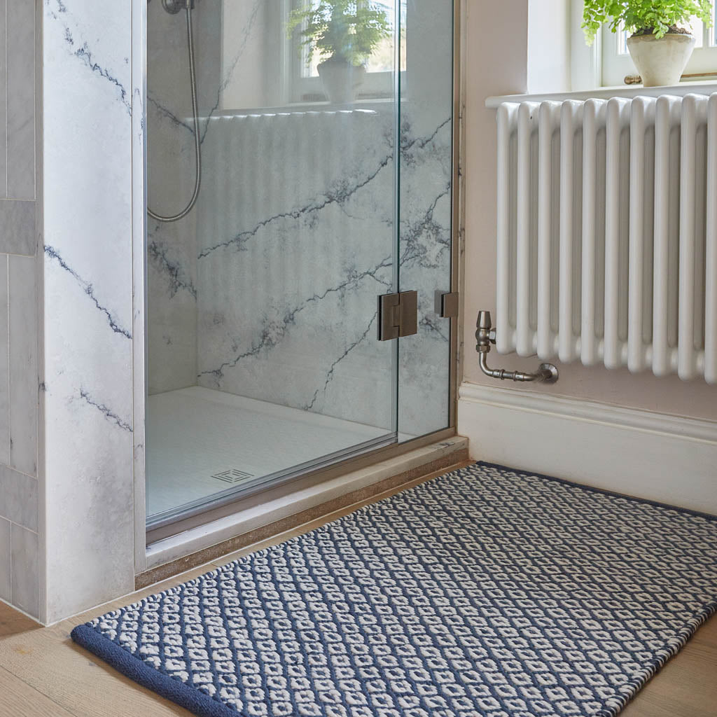 A navy blue rug with a diamond geometric pattern placed on a floor, likely in a bathroom setting given the proximity to a shower door.