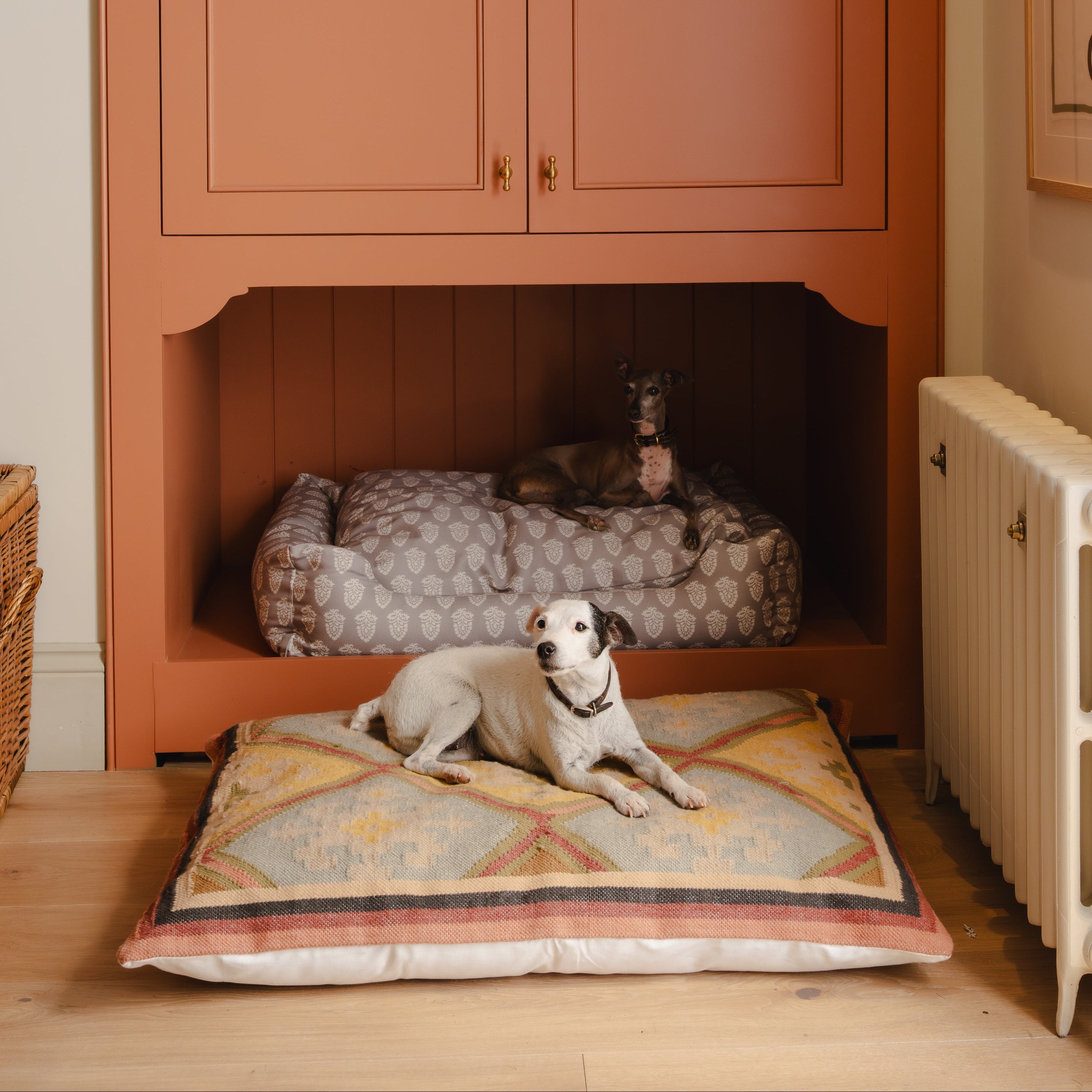 Dog lying on a cushion in front of an arched wooden cabinet with a pet bed inside, in a room with a radiator and framed picture.