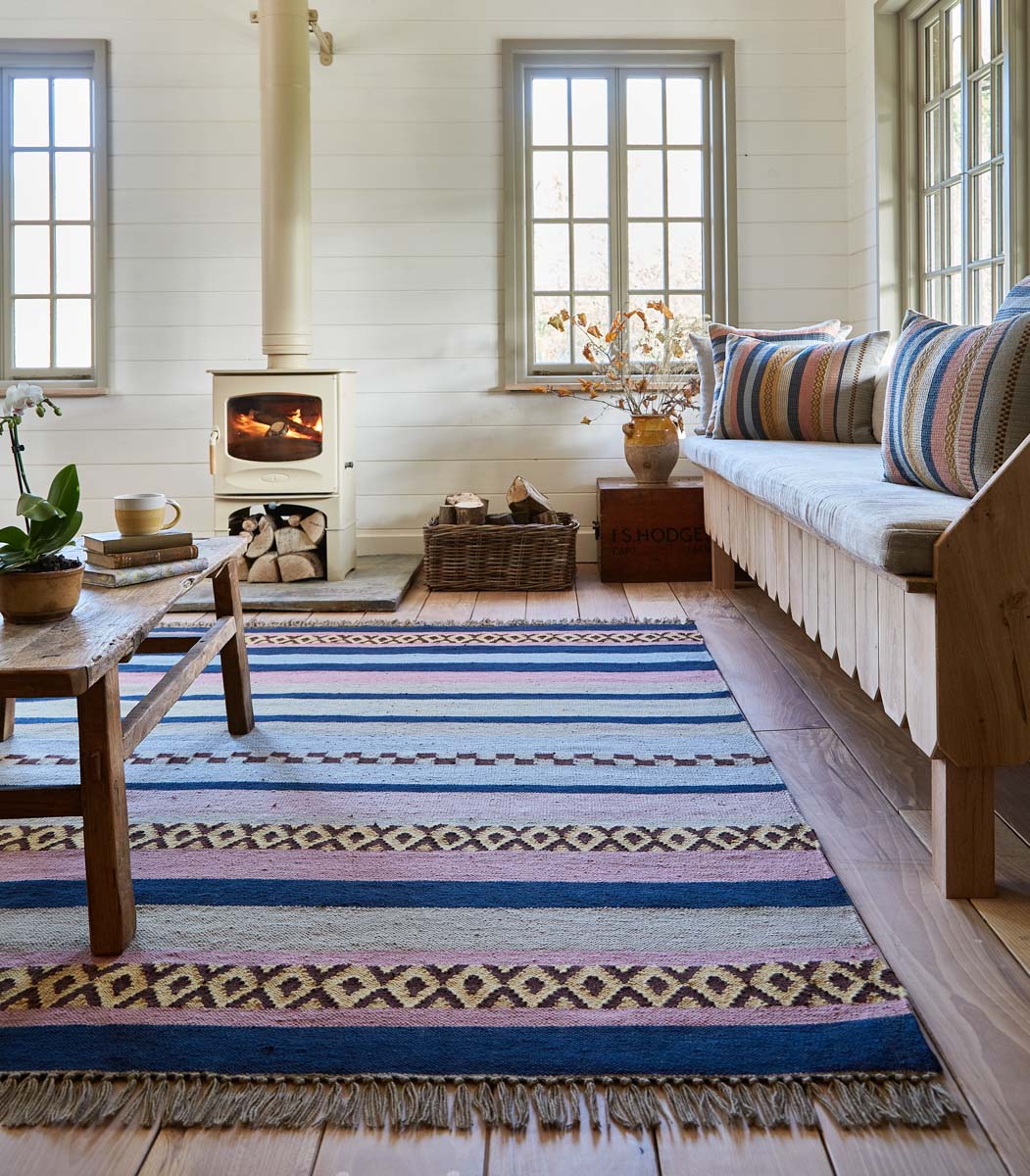 A multicolored geometric patterned rug with stripes and geometric designs in blue, pink, and gold, placed in a room with a wood-burning stove, sofa, and large windows.