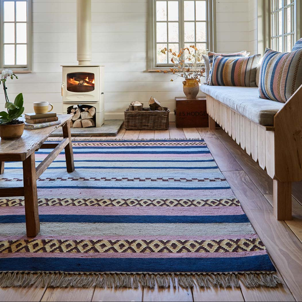 A multicolored geometric patterned rug with stripes and geometric designs in blue, pink, and gold, placed in a room with a wood-burning stove, sofa, and large windows.