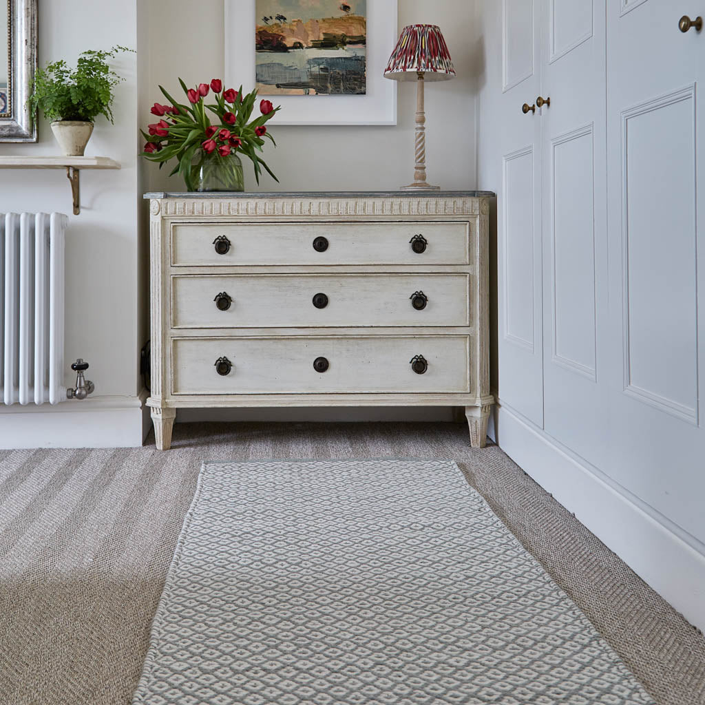 A Dove Grey rug with a subtle geometric diamond pattern placed on a floor in front of a chest of drawers.