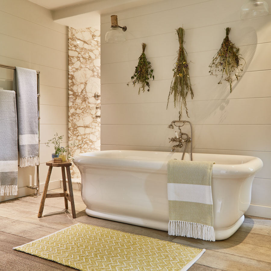 A yellow geometric patterned chenille rug placed on a floor, with a white bathtub and wooden stool in the background, against a wall with a hanging plant.