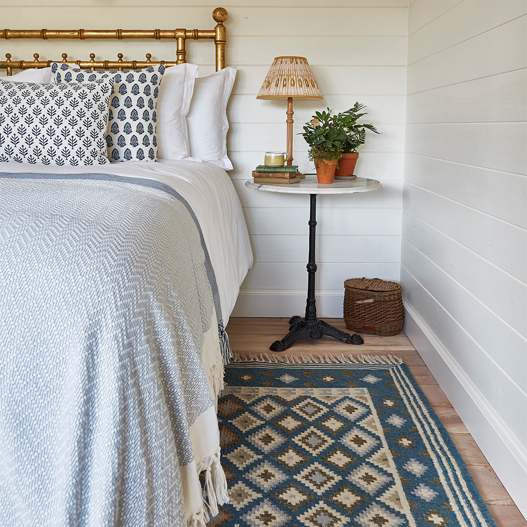 A multicolored geometric patterned rug placed on a floor, presumably a kilim rug, in a room setting with a bed and side table.