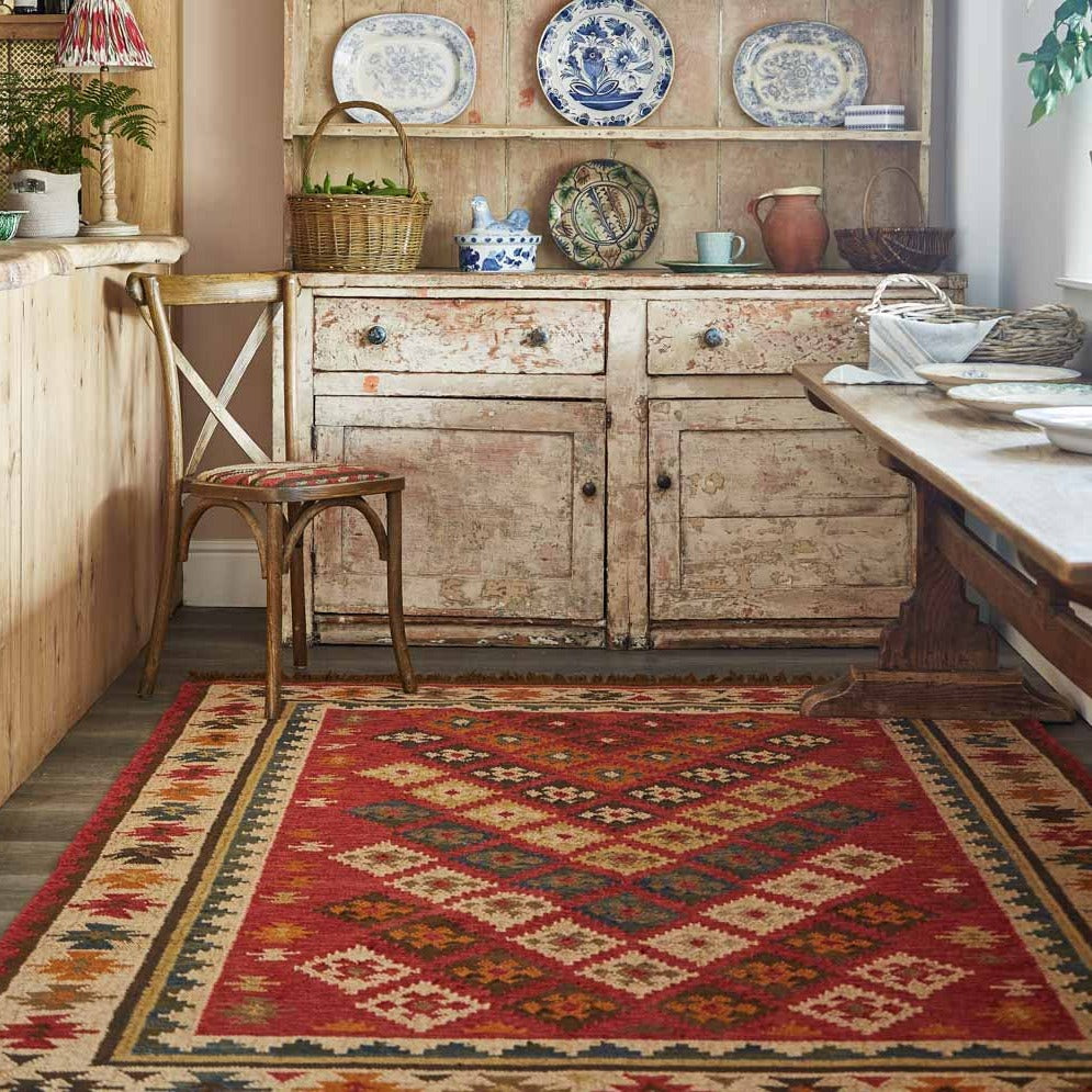 A red multicolored geometric patterned rug placed on a wooden floor within a room setting, featuring furniture and decorative items.