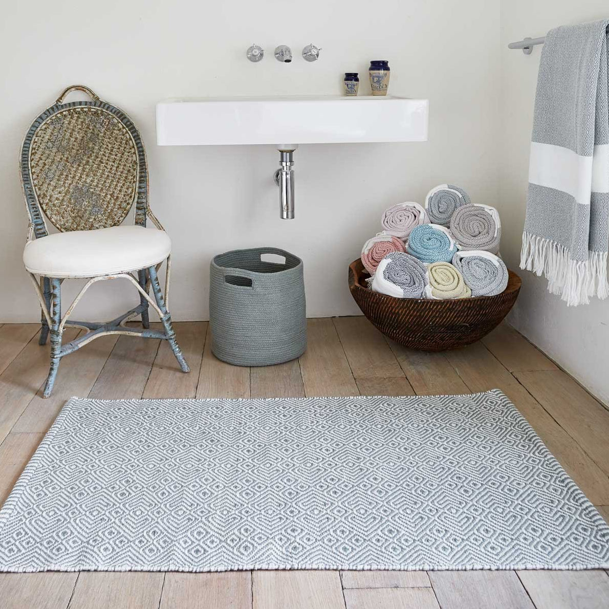 A grey geometric pattern rug placed on a wooden floor in a room with a chair, a bowl of towels, and a shelf with bathroom accessories.