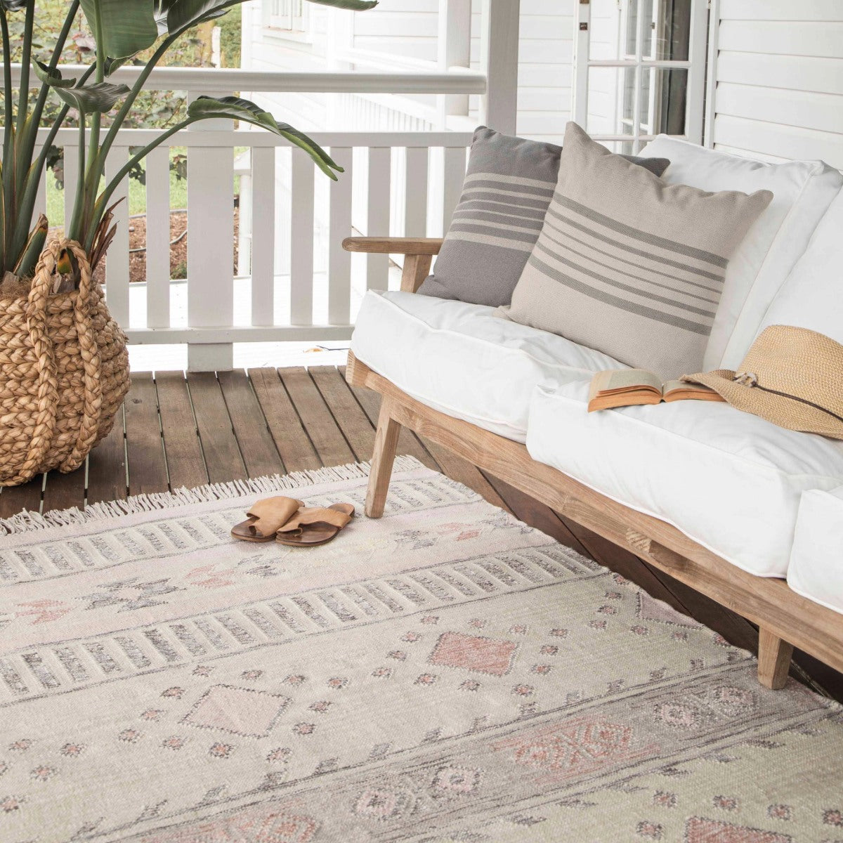 Weaver Green's Andalucia Cadiz Rug in a neutral and pink kilim pattern placed on wooden decking with sandals on top and a sofa.