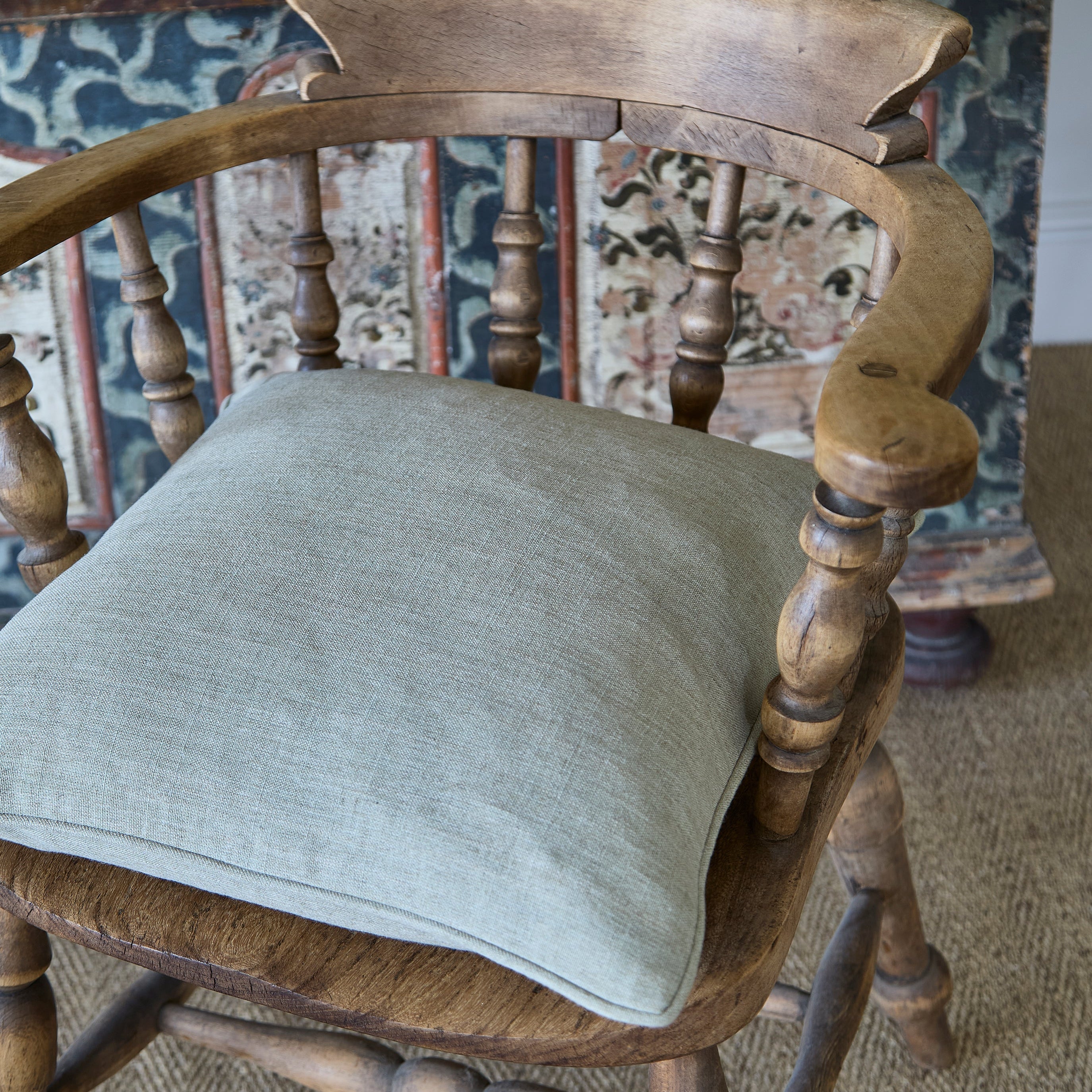 A square light sage green linen throw pillow resting on a rustic wooden armchair with turned spindle legs, set against a background of patterned tapestry and woven rug.