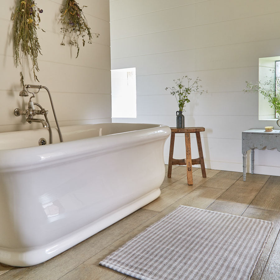 A striped rug in beige and grey colors placed on the floor in front of a bathtub, with a wooden stool and a plant decoration on the wall.