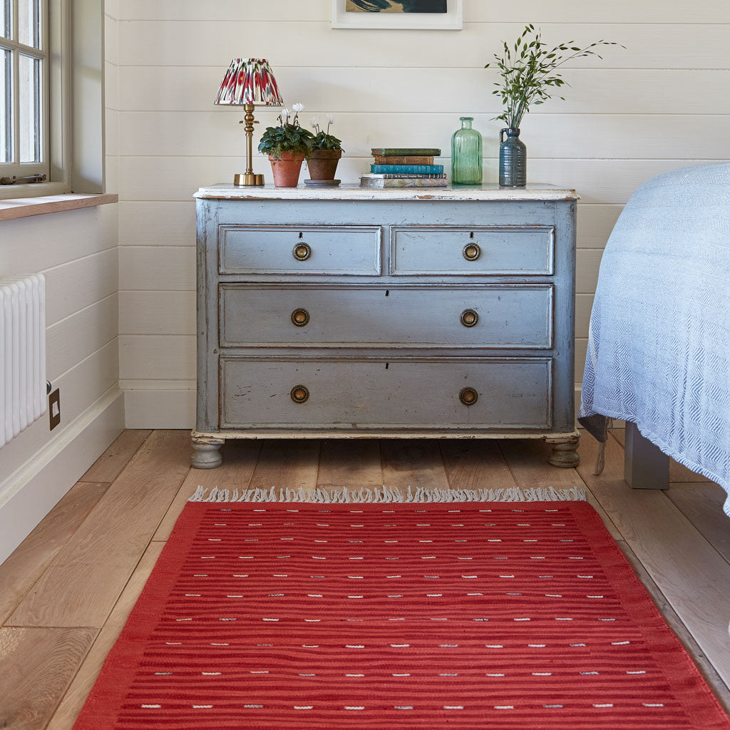A Cosmo Red Rug with a bohemian striped design placed on a floor in front of a dresser with items on top, in a room with a bed and window in the background.