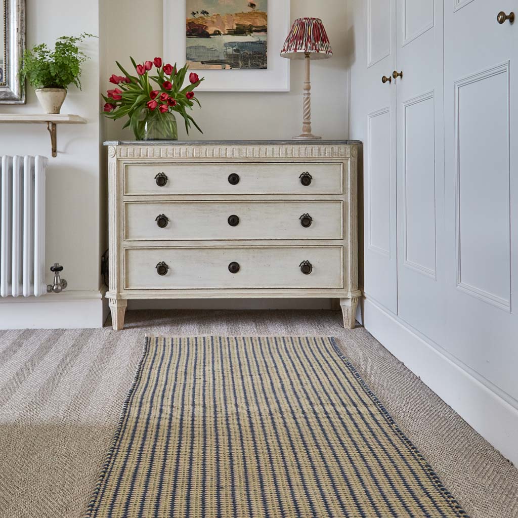 A striped rug in navy blue and gooseberry yellow placed on the floor in front of a chest of drawers, with a plant and a painting on the wall in the background.