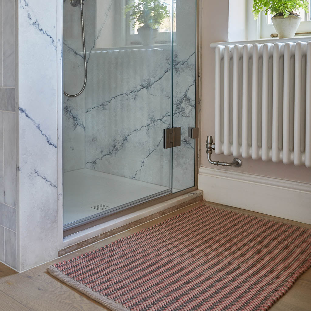 A hand-woven rug with a striped pattern in coral and charcoal colors, placed on a floor with a glass shower enclosure and a radiator in the background.