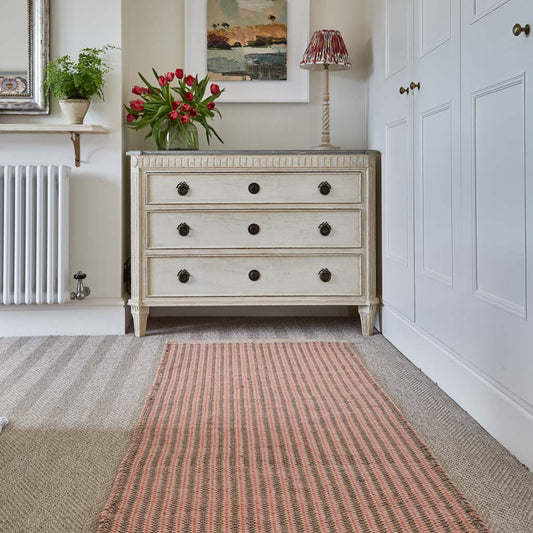 A hand-woven rug with a striped pattern in coral and charcoal colors, placed on a floor with a dresser 