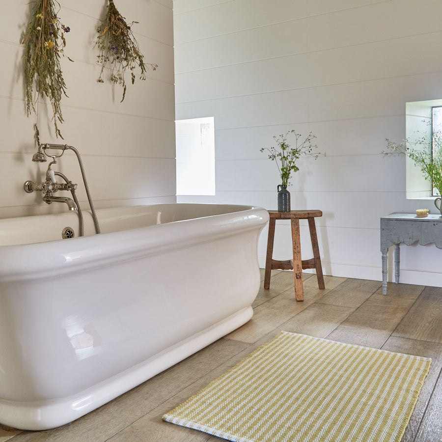 A yellow striped rug placed on a floor, next to a bathtub and a wooden stool with a vase, with a wall-mounted plant decoration above.