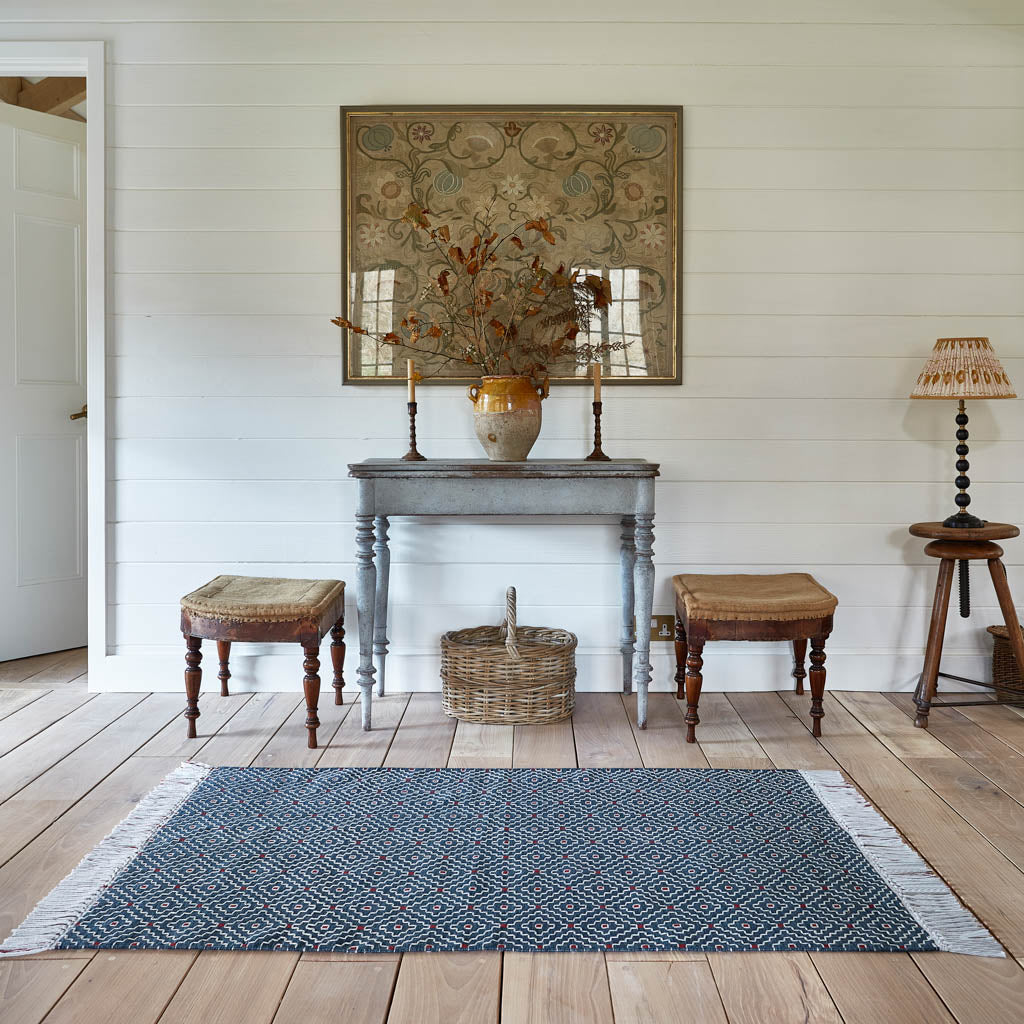 A hand-woven geometric rug with a decorative top-stitched pattern in ecru and red against an indigo blue background, displayed on a wooden floor in a room setting with furniture.