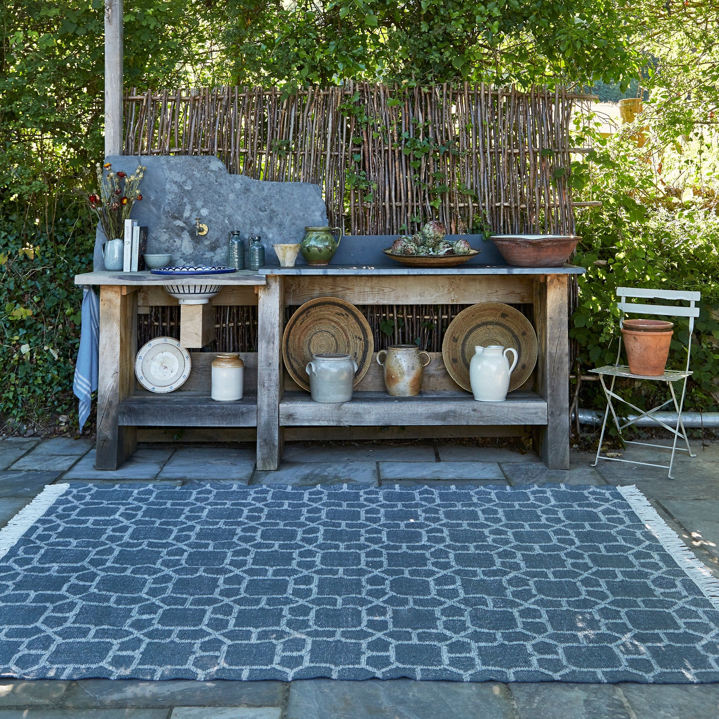 Outdoor kitchen setup with wooden table, pots, and a chair in a garden setting.