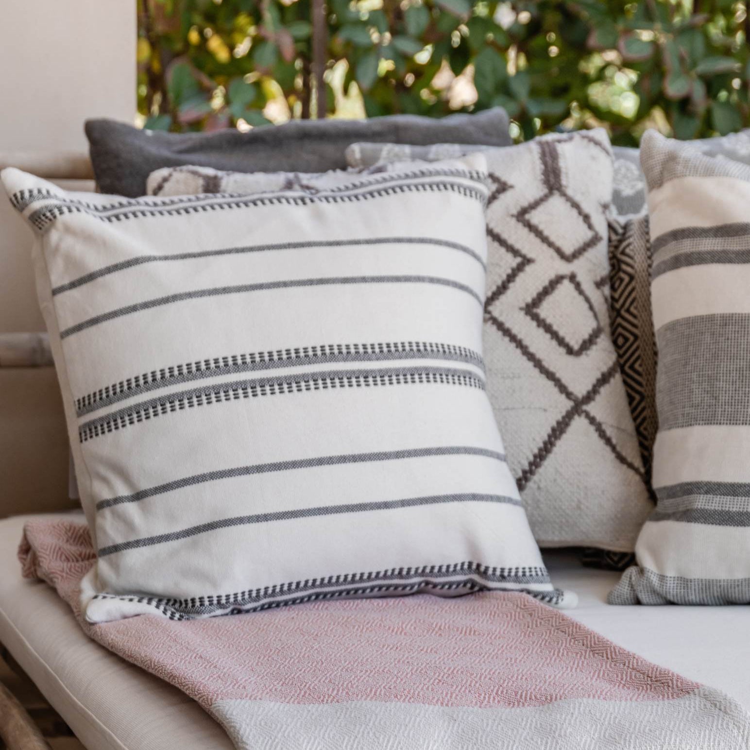 Outdoor setting with a white cushioned bench adorned with patterned cushions and a pink blanket, set against a blurred natural background.