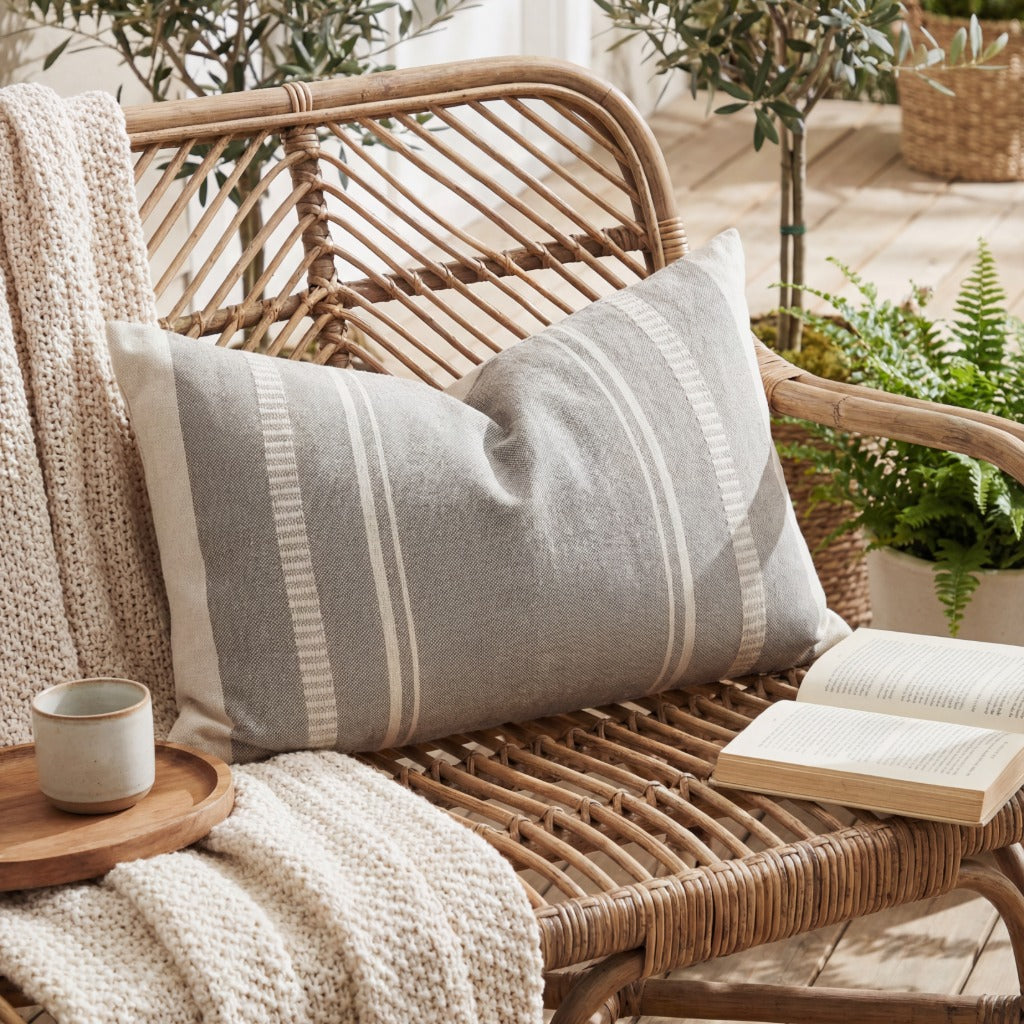 Grey Moli Chinchilla outdoor cushion placed on a wicker chair outside in the sunshine with a book, a blanket and a mug of tea.
