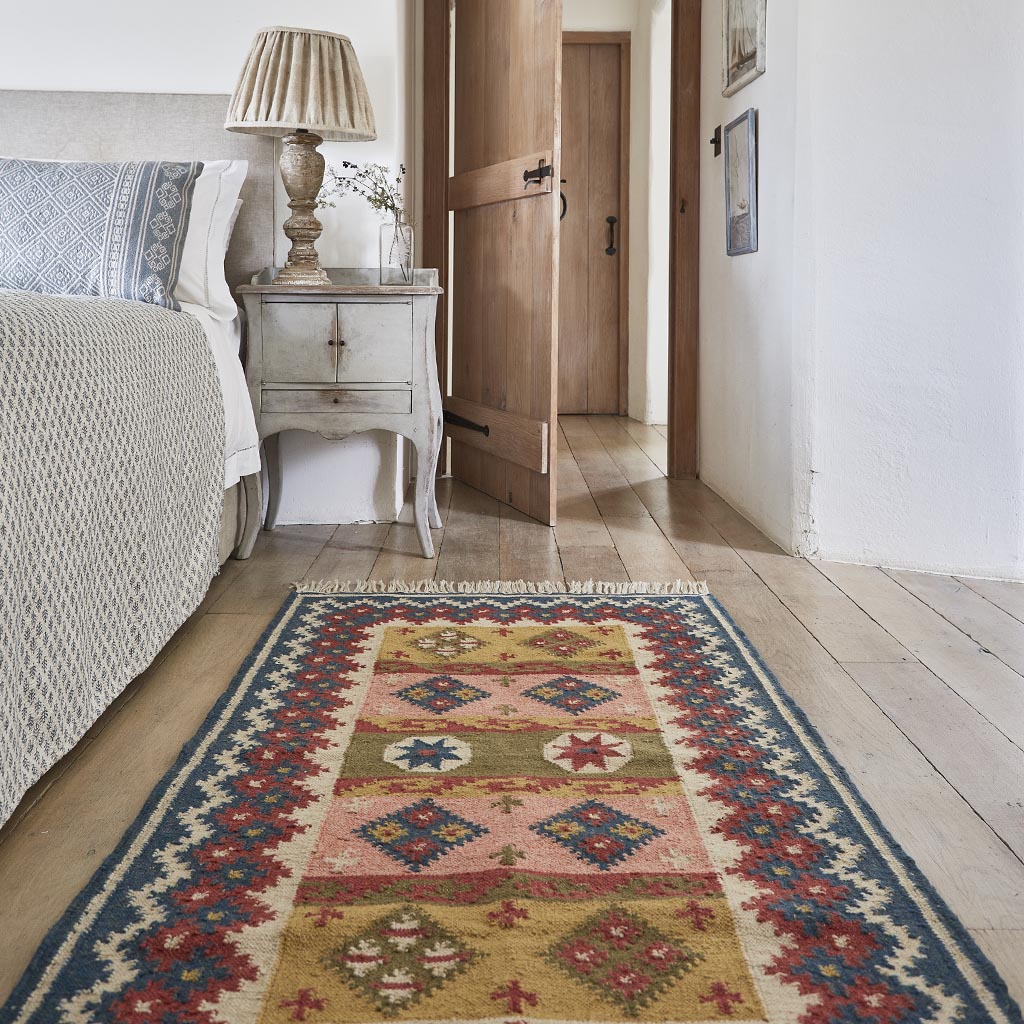 Geometric patterned rug in blue pink red and olive colourways next to a bed