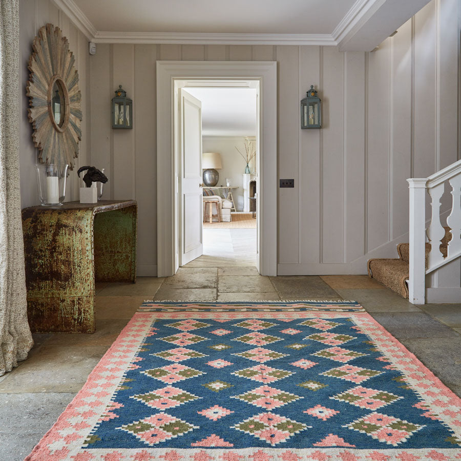 A blue and pink kilim rug with geometric patterns placed on a stone floor in a room with light wooden paneling and decorative wall mirrors.