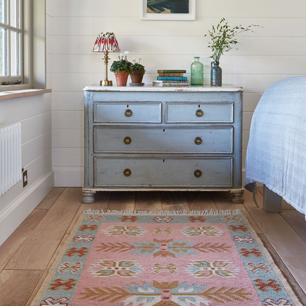 A pink, pale blue and neutral coloured rug at the end of a bed 
