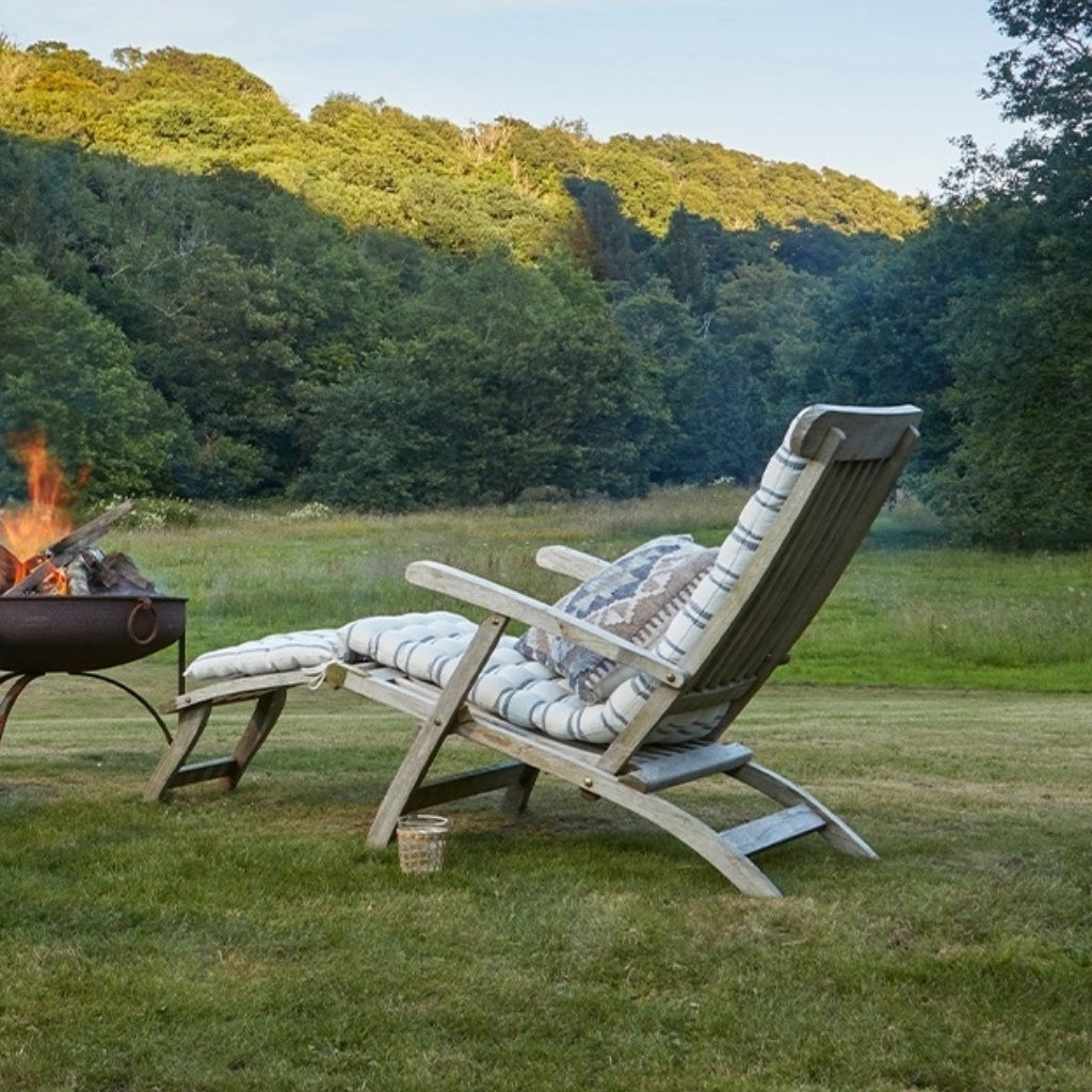 Wooden lounge chair with striped bench cushions and a kilim cushion by a fire pit in a grassy field with trees.