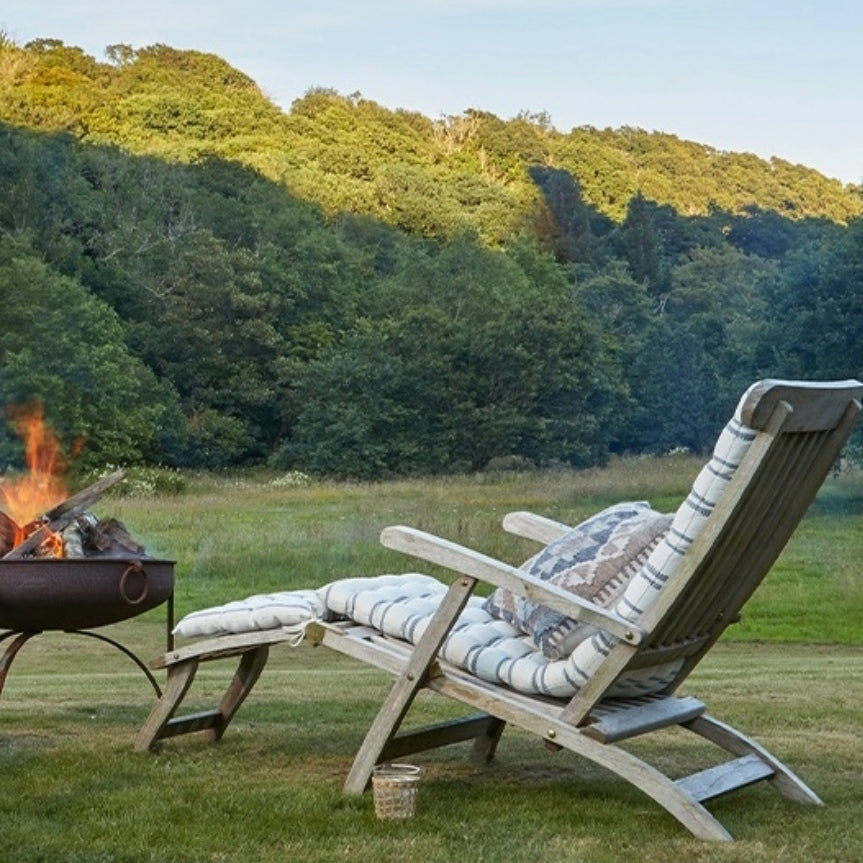 Wooden lounge chair with striped bench cushions and a kilim cushion by a fire pit in a grassy field with trees.