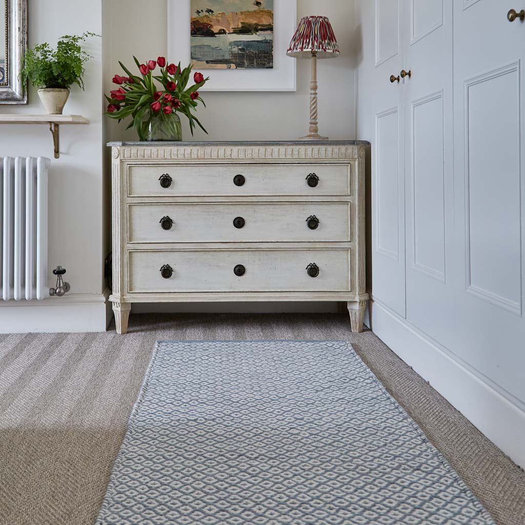 A sky blue geometric rug with a thistle pattern placed on a floor in a room with a wooden cabinet and a floral arrangement.
