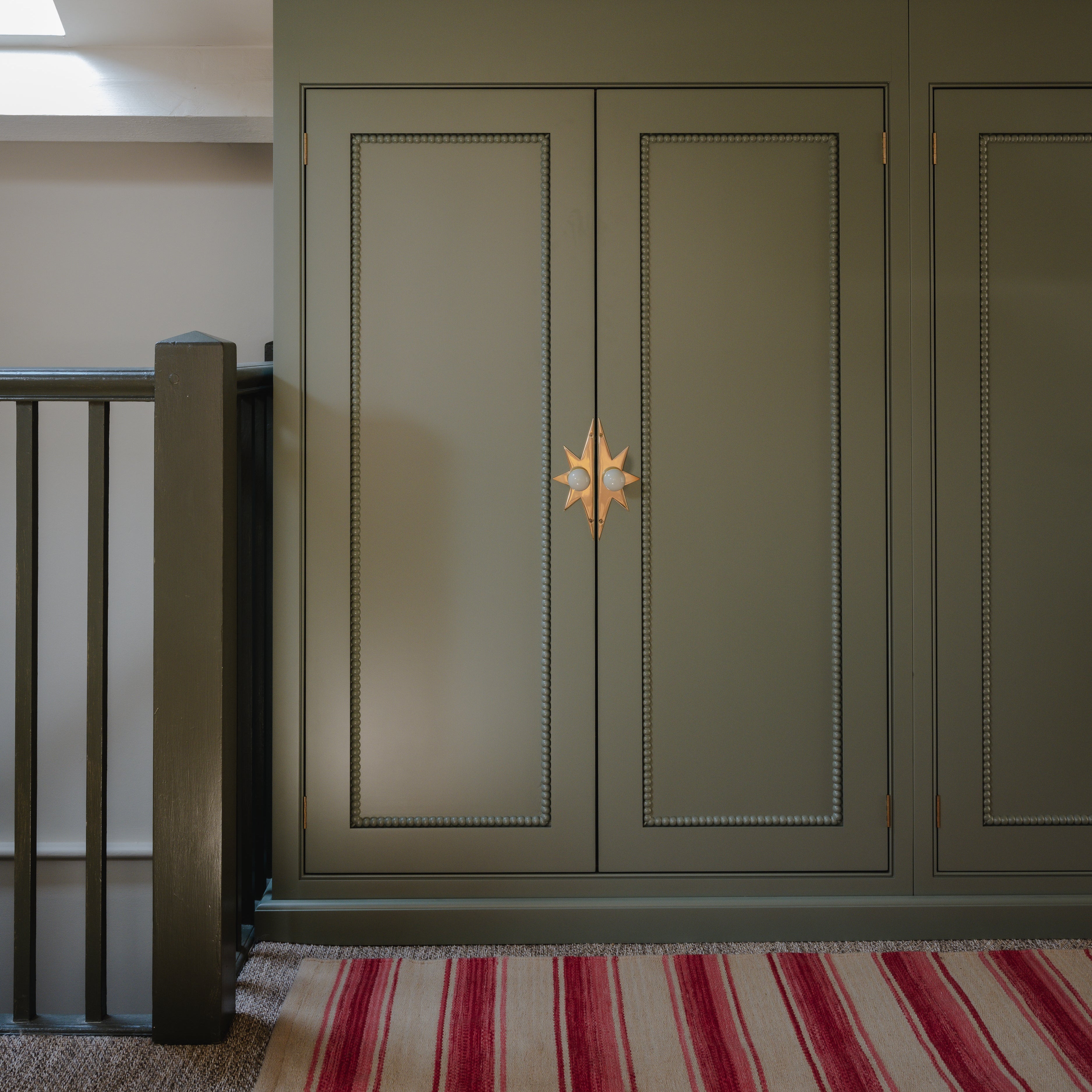 Green cabinet with a decorative handle in a room with a striped red, pink and neutral rug and staircase.