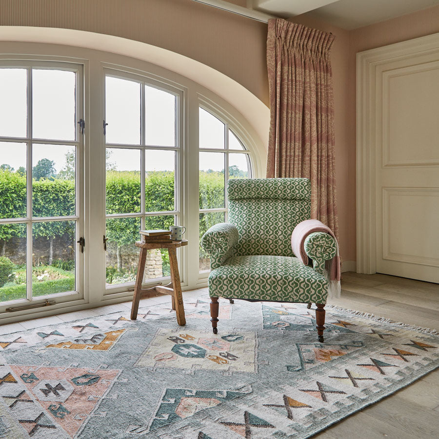 A multicolored geometric patterned rug with blue, beige, and green tones, placed under a green armchair in a room with a view of greenery outside.